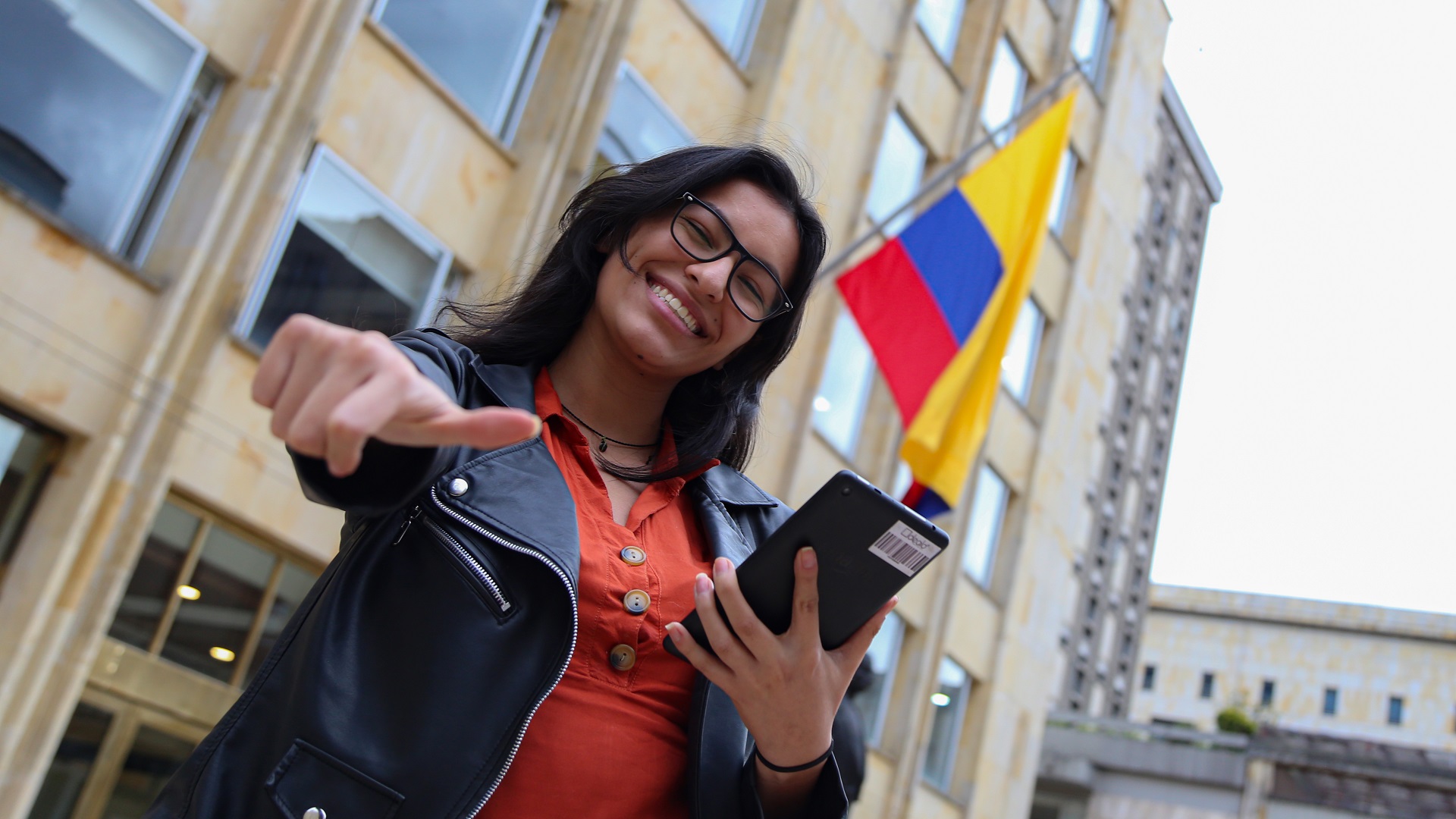 Fotografía: Mujer con celular en la mano y bandera de Colombia en la parte de atrás.